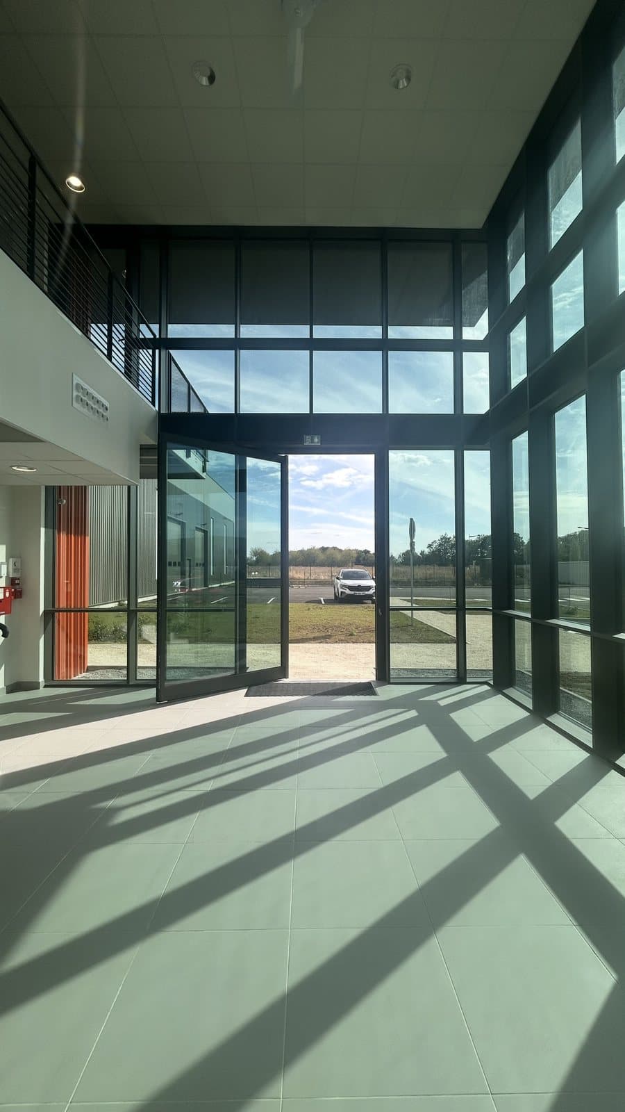 Porte pivot XL ouverte dans le hall, ombres portées de la trame du mur rideau sur le carrelage