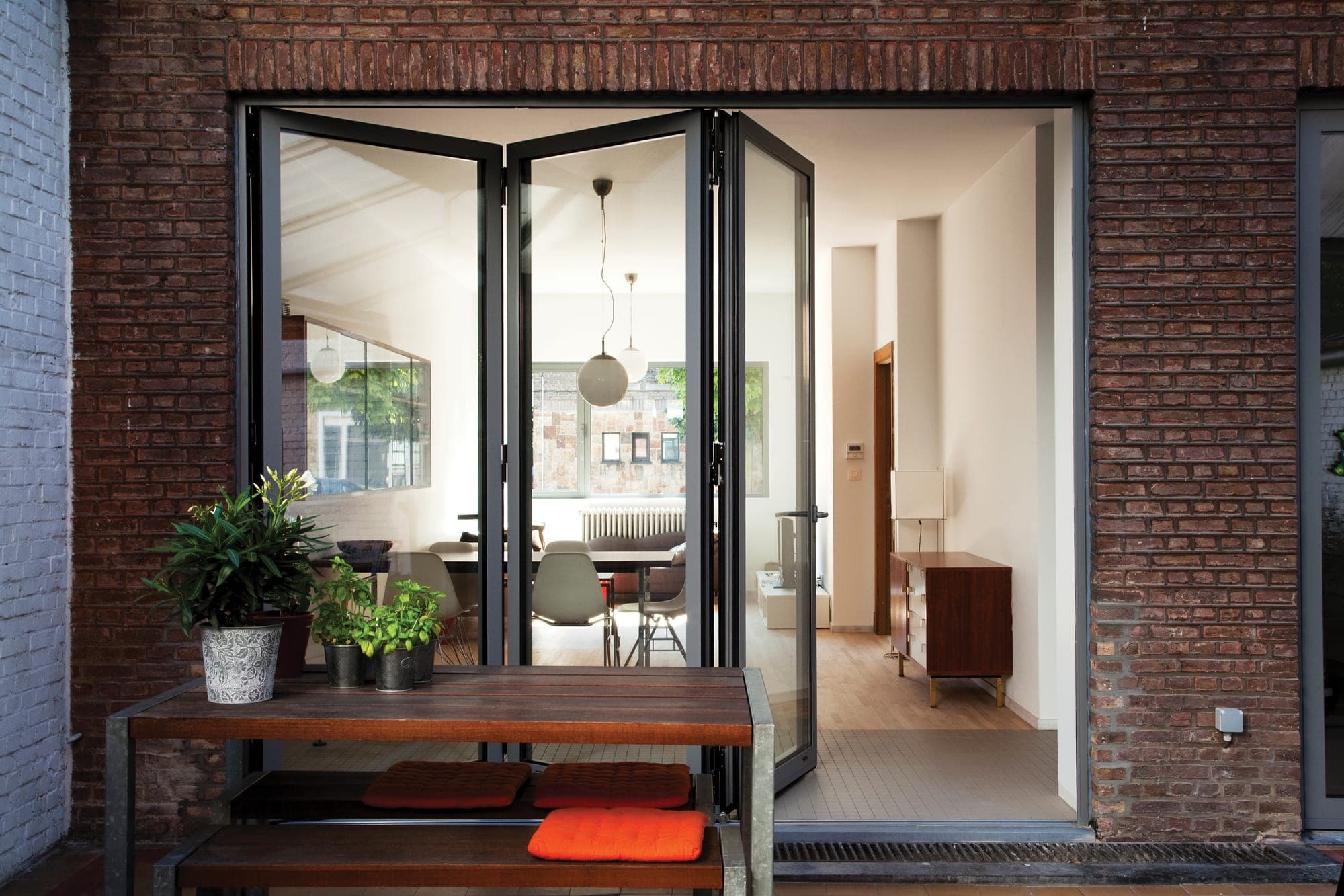 Reynaers CF 77 : porte repliable noire ouverte depuis terrasse avec mur de brique — accès séjour bibliothèque