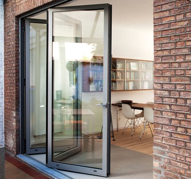 Porte repliable Reynaers CF 77 anthracite ouverte depuis un séjour à murs de brique vers une terrasse, table et bibliothèque